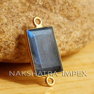 Può includere: Un pendente rettangolare color oro con una gemma blu scuro sfaccettata. Il pendente ha un anello ad ogni estremità per il fissaggio a una catena o ad un altro componente di gioielleria. Il testo "NAKSHATRA IMPEX" è visibile.