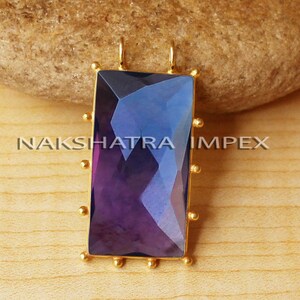 May include: A rectangular pendant with a faceted, color-shifting gemstone in shades of purple and blue, set in a gold-colored frame. The pendant has small, gold-colored accents around the edges. The text "NAKSHATRA IMPEX" is visible.
