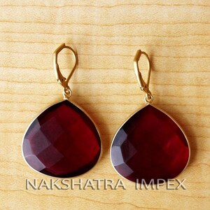May include: A pair of gold earrings with a teardrop-shaped red gemstone. The earrings have a simple design and are perfect for everyday wear.