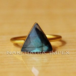 May include: A gold ring with a triangular blue labradorite gemstone. The ring is on a wooden surface.