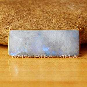 May include: A rectangular, iridescent moonstone pendant with a gold frame. The pendant is set on a wooden surface with a rock behind it. The text "NAKSHATRA IMPEX" is visible on the surface.