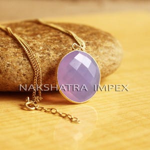 May include: A gold chain necklace with a lavender gemstone pendant. The pendant is oval shaped and has a faceted design. The necklace is on a brown rock.