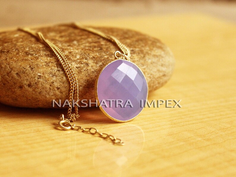 May include: A gold chain necklace with a lavender gemstone pendant. The pendant is oval shaped and has a faceted design. The necklace is on a brown rock.