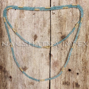 Puede incluir: Un collar de doble cadena hecho de cuentas de color azul claro con detalles dorados. El collar est&aacute; sobre una superficie de madera.