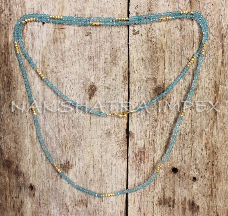 Puede incluir: Un collar de doble cadena hecho de cuentas de color azul claro con detalles dorados. El collar est&aacute; sobre una superficie de madera.
