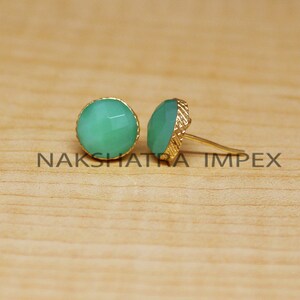 May include: A pair of gold stud earrings with green gemstone accents. The earrings are on a wooden surface.