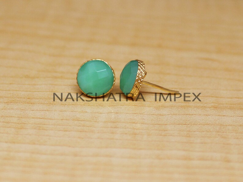 May include: A pair of gold stud earrings with green gemstone accents. The earrings are on a wooden surface.