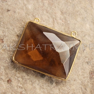 Puede incluir: Un colgante dorado con una gran piedra preciosa marrón facetada. El colgante tiene dos anillos de salto para sujetarlo a una cadena o cordón.