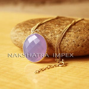 May include: A gold chain necklace with a purple oval pendant. The pendant has a faceted design and is set in a gold frame. The necklace is on a brown rock.