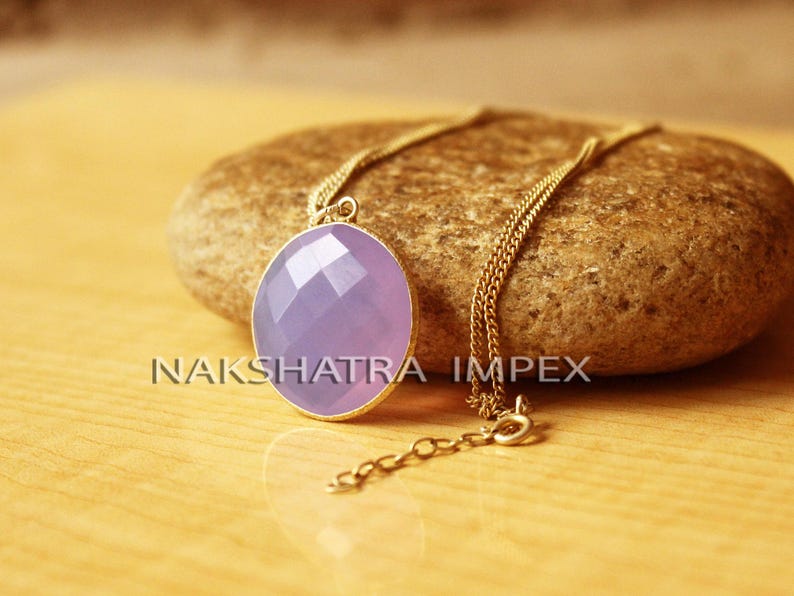 May include: A gold chain necklace with a purple oval pendant. The pendant has a faceted design and is set in a gold frame. The necklace is on a brown rock.