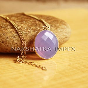 May include: A gold chain necklace with a purple gemstone pendant. The pendant is oval shaped and has a faceted design. The necklace is resting on a brown rock.