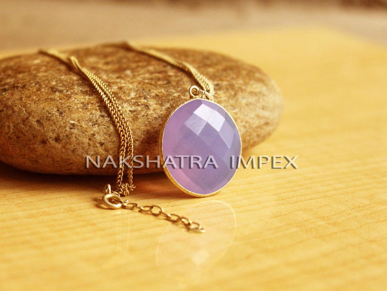 May include: A gold chain necklace with a purple gemstone pendant. The pendant is oval shaped and has a faceted design. The necklace is resting on a brown rock.