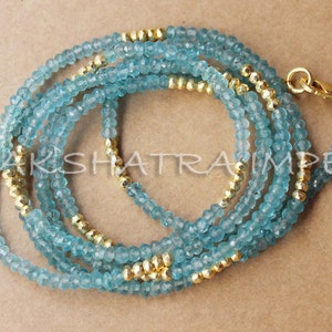 Puede incluir: Un collar de capas hecho de cuentas de color azul claro y dorado. El collar est&aacute; hecho de m&uacute;ltiples hebras de cuentas, con las cuentas doradas intercaladas entre las cuentas azules.