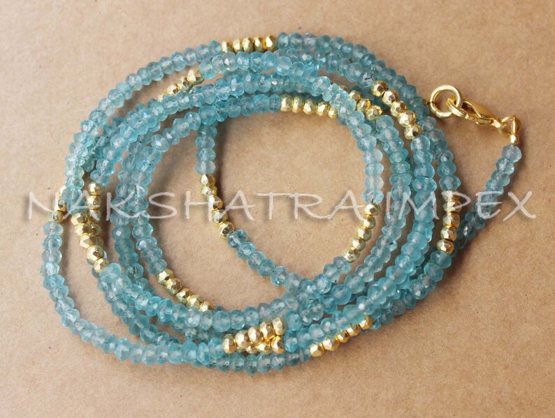 Puede incluir: Un collar de capas hecho de cuentas de color azul claro y dorado. El collar est&aacute; hecho de m&uacute;ltiples hebras de cuentas, con las cuentas doradas intercaladas entre las cuentas azules.