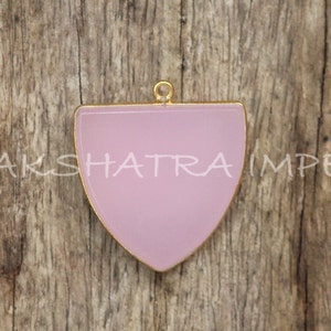 Pode incluir: Um pingente semi-circular de cor rosa pálido com uma moldura de metal dourado e um laço na parte superior para pendurar. O pingente está sobre uma superfície de madeira.