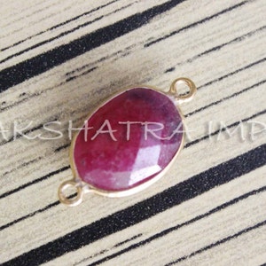 Może przedstawiać: Złoty owalny łącznik z czerwonym kamieniem szlachetnym. Łącznik ma po dwie pętle z każdej strony.