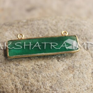 May include: A gold-plated rectangular pendant with a green gemstone. The pendant has two jump rings for attaching to a chain or cord.