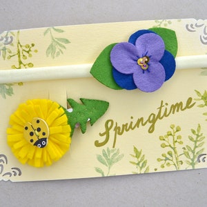 May include: Two felt flower hair ties on a yellow card with the word "Springtime" written in gold. One hair tie features a yellow flower with a ladybug and the other features a purple flower with green leaves.