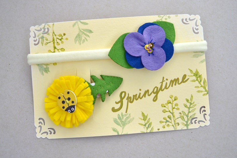 May include: Two felt flower hair ties on a yellow card with the word "Springtime" written in gold. One hair tie features a yellow flower with a ladybug and the other features a purple flower with green leaves.