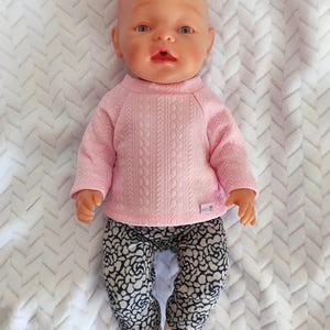 Può includere: Un outfit per una bambola bebé composto da una maglietta rosa a maniche lunghe con un motivo a treccia e leggings con stampa floreale in bianco e nero.