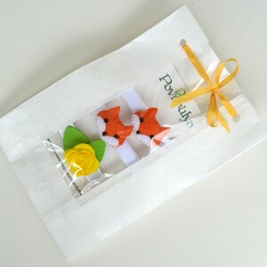 May include: A white paper bag with a yellow ribbon tied around it. The bag contains two orange fox hair clips and a yellow flower hair clip. The bag has the text "Povižuj" printed on it.