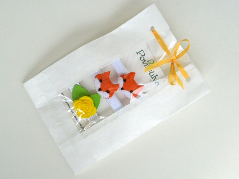 May include: A white paper bag with a yellow ribbon tied around it. The bag contains two orange fox hair clips and a yellow flower hair clip. The bag has the text "Povižuj" printed on it.