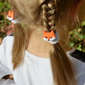 May include: A young girl with blonde hair wearing a white shirt with black lace on the sleeve. Her hair is pulled back into two pigtails with orange fox-shaped hair ties.