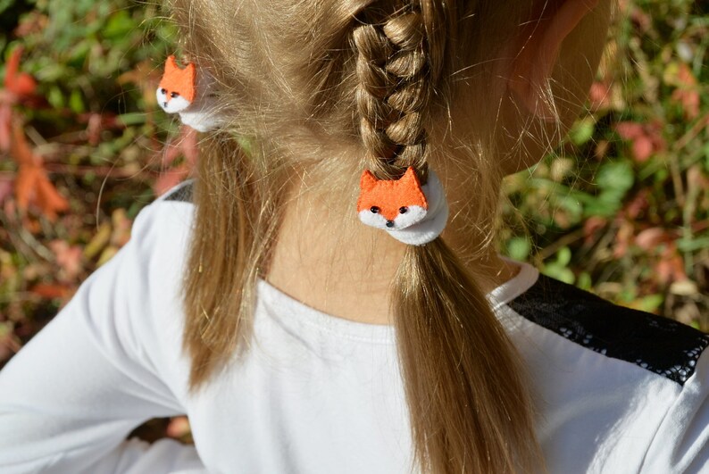May include: A young girl with blonde hair wearing a white shirt with black lace on the sleeve. Her hair is pulled back into two pigtails with orange fox-shaped hair ties.