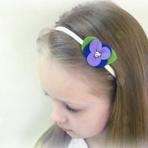 May include: A white headband with a purple, blue, and green felt flower accent. The flower has a gold center.