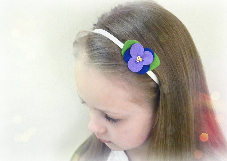 May include: A white headband with a purple, blue, and green felt flower accent. The flower has a gold center.