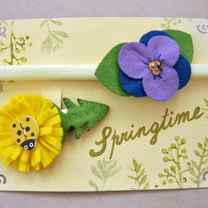 May include: Two felt flower hair clips on a yellow card with the word "Springtime" written in gold script. One clip features a yellow dandelion with a ladybug and the other features a purple flower with a blue center.