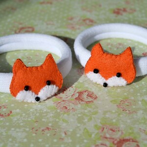 May include: Two white hair ties with orange felt fox faces. Each fox has black eyes and a white nose.