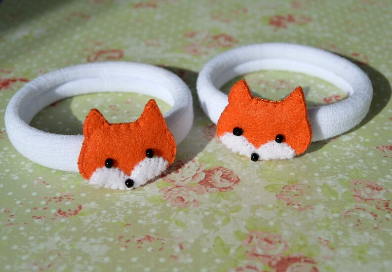 May include: Two white hair ties with orange felt fox faces. Each fox has black eyes and a white nose.