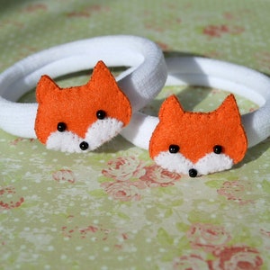 May include: Two white elastic hair ties with orange felt fox head embellishments. Each fox has black eyes and a white nose.