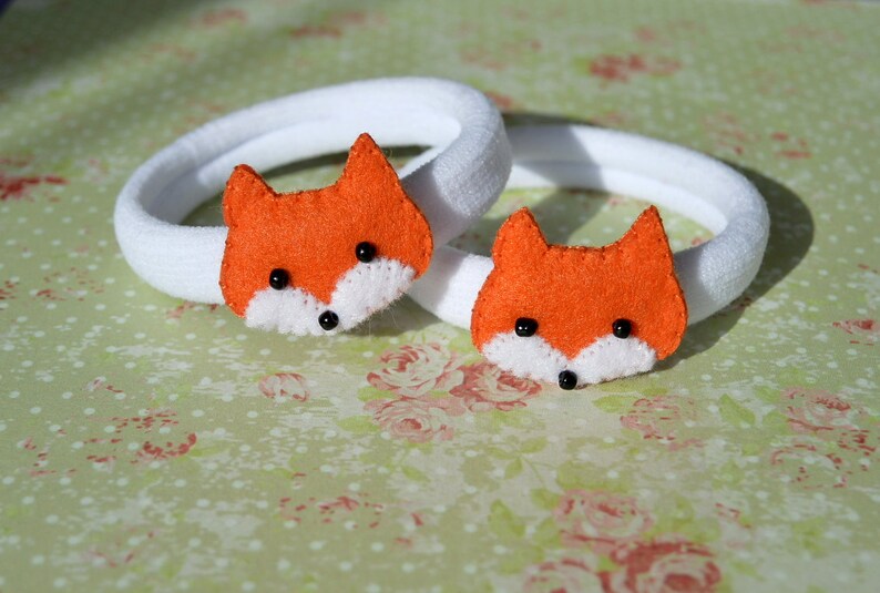 May include: Two white elastic hair ties with orange felt fox head embellishments. Each fox has black eyes and a white nose.