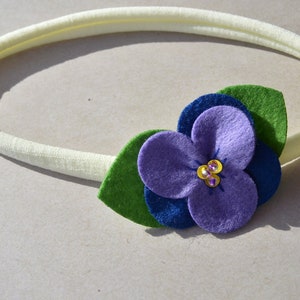 May include: A white elastic headband with a purple felt flower and green felt leaves. The flower has a blue felt center and is decorated with small, sparkly gems.