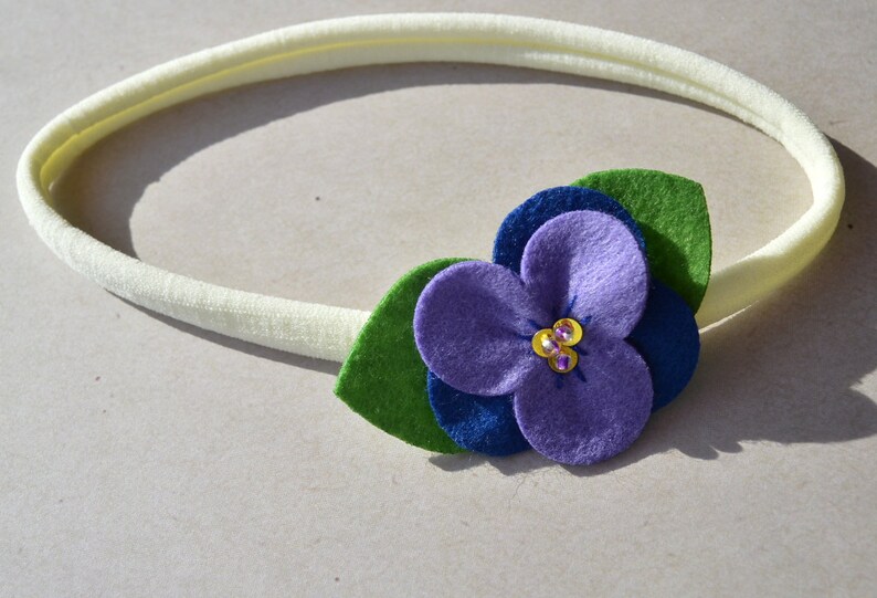 May include: A white elastic headband with a purple felt flower and green felt leaves. The flower has a blue felt center and is decorated with small, sparkly gems.