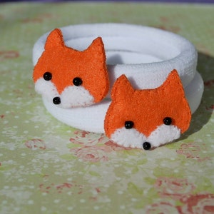 May include: Two white fabric hair ties with orange felt fox faces attached. The foxes have black eyes and white noses.