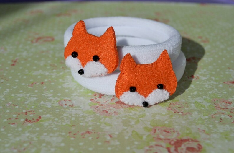 May include: Two white fabric hair ties with orange felt fox faces attached. The foxes have black eyes and white noses.