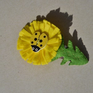 May include: A yellow felt dandelion flower with a green felt stem and a ladybug button in the center.