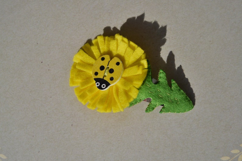 May include: A yellow felt dandelion flower with a green felt stem and a ladybug button in the center.