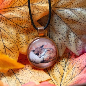 Könnte beinhalten: Eine runde Glas-Anhänger-Halskette mit einem schwarzen Band. Der Anhänger zeigt eine Aquarellmalerei eines schlafenden Fuchses. Der Anhänger ist vor einem Hintergrund aus Herbstblättern platziert.