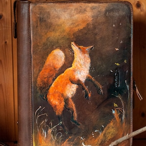 Könnte beinhalten: Ein braunes Lederjournal mit einem lebendigen Gemälde eines springenden orangefarbenen Fuchses. Das Kunstwerk zeigt eine detaillierte Darstellung des Fuchses vor einem dunklen, atmosphärischen Hintergrund mit gelben und weißen Akzenten. Das Journal hat einen sicheren Verschluss.