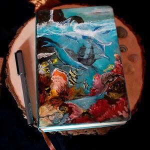 Könnte beinhalten: Ein blaues Notizbuch mit einem bemalten Umschlag, der eine Unterwasserszene darstellt. Der Umschlag zeigt ein Schiffswrack, Korallenriffe, Fische und einen Wal. Das Notizbuch ist geschlossen und liegt auf einer Holzoberfläche.