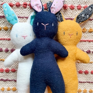May include: Three handmade bunny dolls in white, navy blue, and yellow. Each plush bunny has long ears with patterned fabric lining, embroidered facial features, and a simple body shape. The bunnies are standing together on a patterned surface.