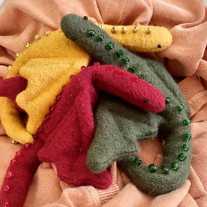 Peut inclure: Gros plan sur des jouets dragons en feutre faits à la main, dans les tons de rouge, jaune et vert. Chaque dragon est orné d'accents de perles sur les ailes et le corps. Les jouets sont disposés sur un tissu pêche, mettant en valeur une esthétique artisanale et fantaisiste.