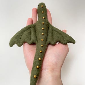 Puede incluir: Un adorno de dragón de fieltro en color verde oliva con dos alas extendidas y una cola larga. El dragón está decorado con una fila de pequeñas cuentas doradas metálicas que recorren el centro de la espalda y la cola, con dos cuentas negras diminutas como ojos.
