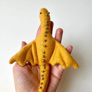 May include: A mustard-yellow felt dragon toy with outstretched wings. The dragon has small black bead eyes and is decorated with gold-coloured beads along its spine. The wings have subtle stitching details.