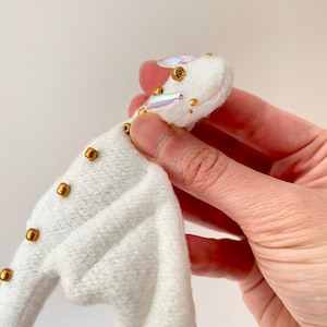May include: A white felt dragon figurine featuring iridescent, oval-shaped accents on the head and small, metallic gold beads arranged along the spine and face. The creature has a soft, textured surface and is crafted with detailed stitching.