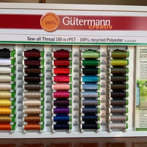 May include: A display of Gutermann creativ Sew-all Thread spools in various colors. The spools are arranged in rows on a white background. The text "Sew-all Thread 100 m rPET - 100% recycled Polyester" is displayed at the top of the display.